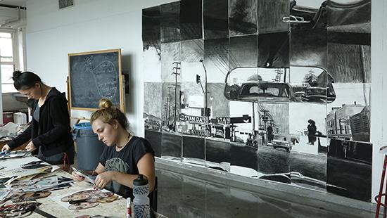 Two women working at a table with a large black-and-white collage on the wall
