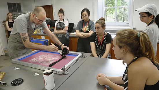 man adding paint to a print screening apparatus while students watch