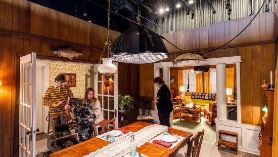 People on a filmset adjusting a film camera in a dining room scene.