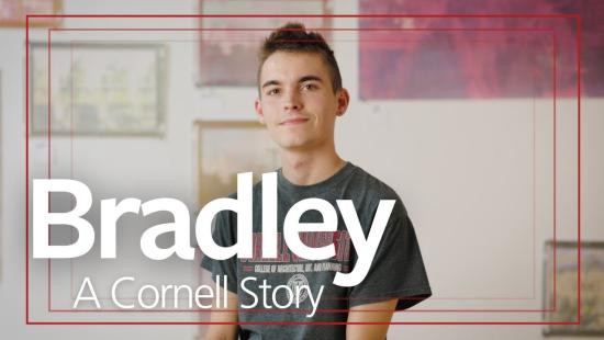 A person wearing a grey t-shirt sitting in front of a wall of hanging art work.