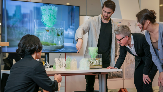 Four people observing a physical model on a table with a large television screen in the background.