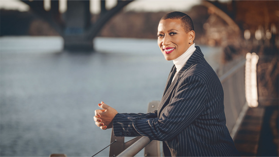 a black woman with short hair and a pin striped blazer leans on a railing annd smiles.