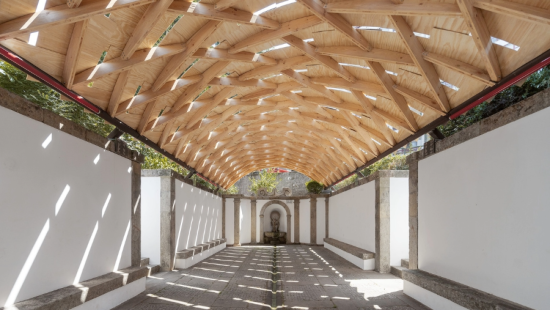 Timber vault roof over restored stone ruin in Braga, casting patterned light and creating a community gathering space.