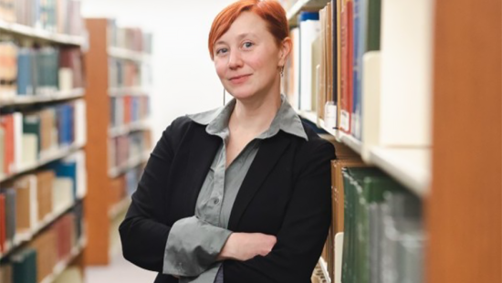 Woman with short red hair and wearing a black suit jacket leaning against a bookshelf in a library