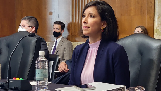 Woman in a blue suit seated at a table in front of a microphone with spectators gathered behind her