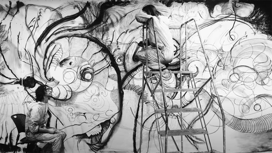 Two men wearing white coveralls, one seated and one on a ladder, drawing on the wall