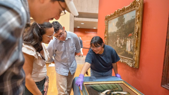 People stand around a painting set on a table in a museum.