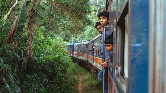 three Indian males standing at the open door of a moving train through the countryside