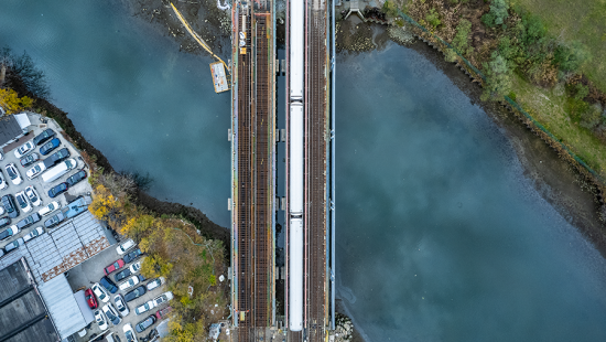 Aerial view of a train bridge over a river. Clouds are reflected in the water. In the upper right corner is the edge of land with green grass and trees. In the bottom left corner is land with some trees and about twenty cars and an industrial building.