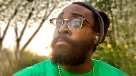 A person wearing a black and grey beanie and glasses with a gold necklace and green shirt.