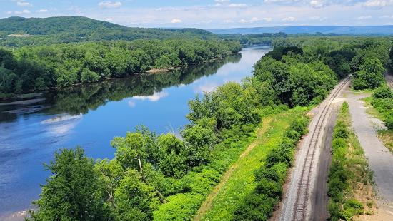 River with railroad tracks beside along trees and hills with a partly cloudy sky.