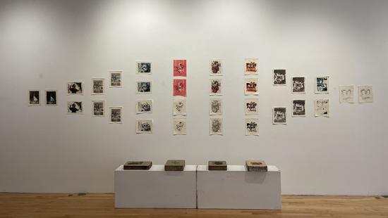 A view of a gallery exhibition, with numerous small prints in a grid on the wall and two white pedestals in front, each with two book-like objects on top.