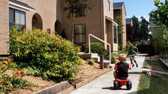 Two kids playing on a sidewalk with a person standing on a porch watching them.