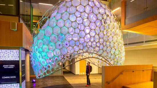 A person looks up from under a colorful, dome-shaped lattice structure in a gallery space.