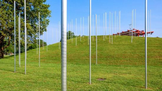 A series of large silver poles protruding from a grassy field with a red structure in the distance.
