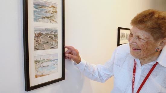 A person with a white shirt and red cornell university lanyard pointing at a frame willed with watercolor paintings.