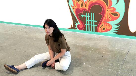 Person sitting on a concrete floor, a brown green red and orange mural painted on a wall.