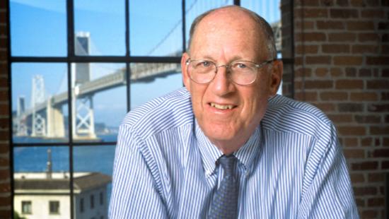 A person with balding head wearing glasses, window showing a suspension bridge over water. 