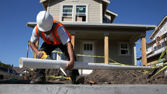 Construction worker in white hard hat laying pipe in front of a partially constructed house.