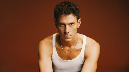 Man with short dark curly hair wearing a sleeveless undershirt and silver chain against a brown background.