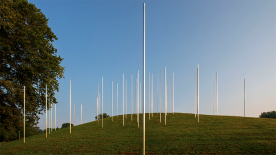 58 vertical metal poles set on a grassy mound, blue sky.