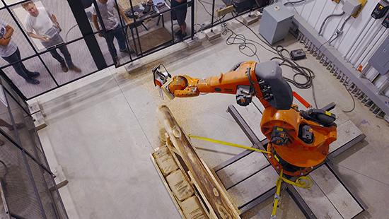 men watch a robotic arm shaping wood