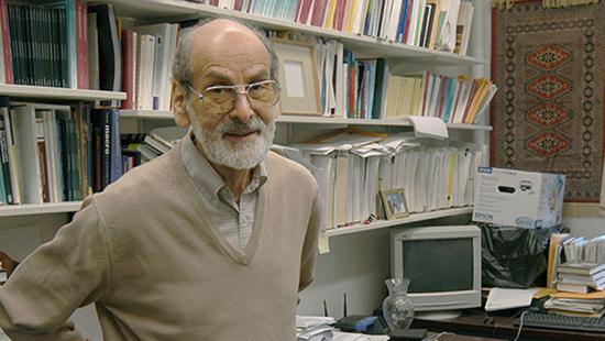 A bearded man wearing glasses stands near bookshelves and a computer