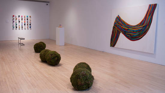 Gallery space with six large moss balls on the floor and brightly colored paintings on the walls