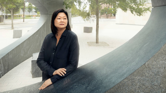 Person with shoulder-length black hair wearing black blouse, hand resting on concrete sculpture