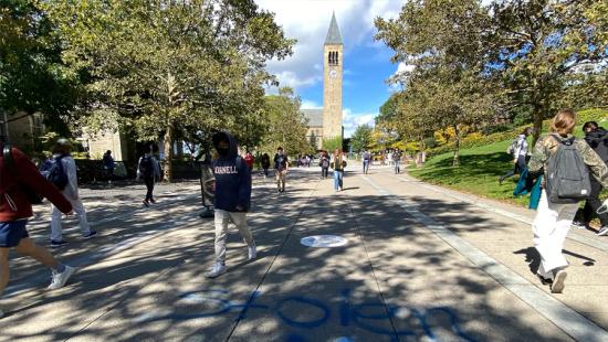 People walking on a paved plaza with a bell tower and trees in the background.