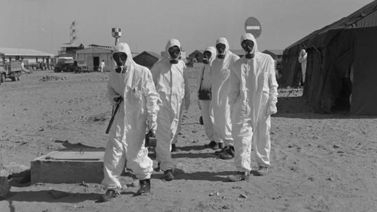 Five workers in protective white clothing and gas masks walking across sand with tents and buildings in background