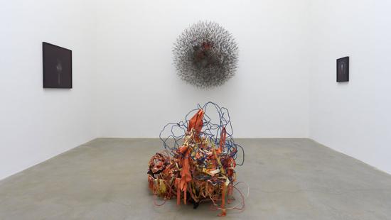 sculpture made of blue wire and orange and yellow ribbon at center, dark canvases and dark wire sculpture at back