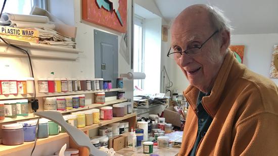A person in an art studio with shelves of paint and supplies.
