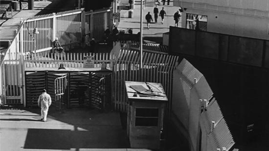 People walking around tall metal fencing and concrete buildings.