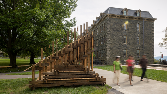 People walking along a sidewalk passing a wooden sculpture, large stone building at back.