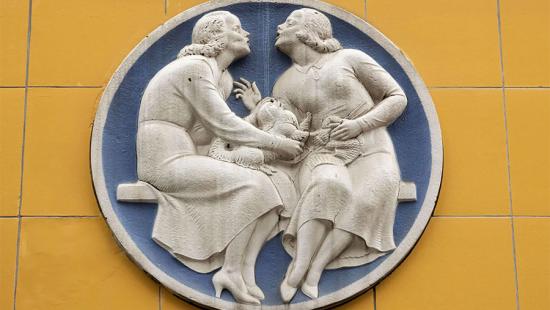white and blue terra-cotta sculpture showing two women seated on a bench with dog in between
