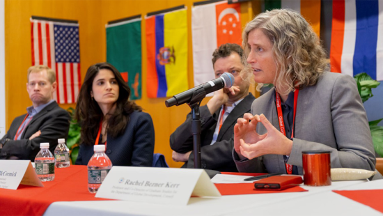 Woman seated at a table speaking into a microphone while three other people look on.