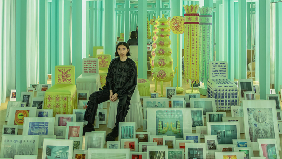 Man dressed in black surrounded by framed photographs and yellow sculptures.