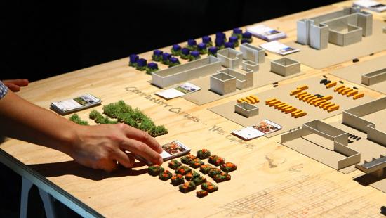 a hand moves around urban scale model pieces on a wood table