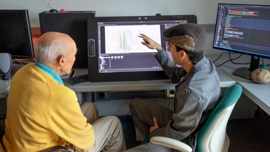 Two people seated, one gesturing at the computer screen behind.