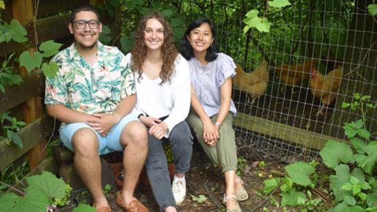 Three people seated in a leafy arbor with chickens behind a wire fence.