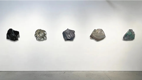 A series of large rocklike formations hanging on a white gallery wall with a concrete floor beneath.
