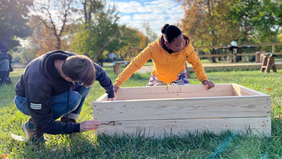 Two people crouched around a large wooden container in the middle of a lawn.