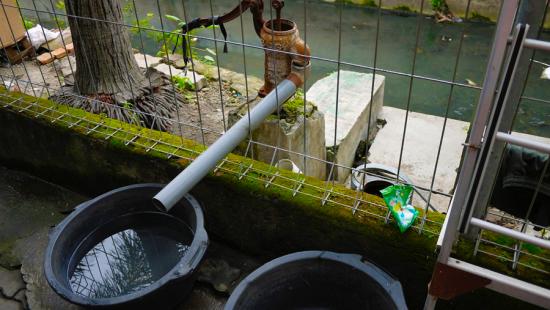 Two black buckets underneath a water spout connected to a small stream.