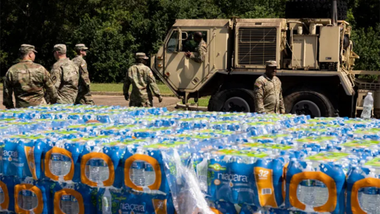 Pallets of water with soldiers and heavy military vehicle behind