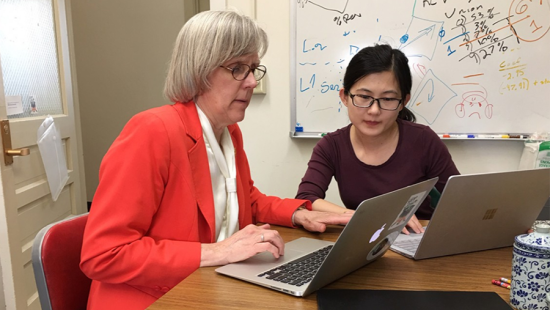 Two people at a table in an office, each with silver laptop, in discussion