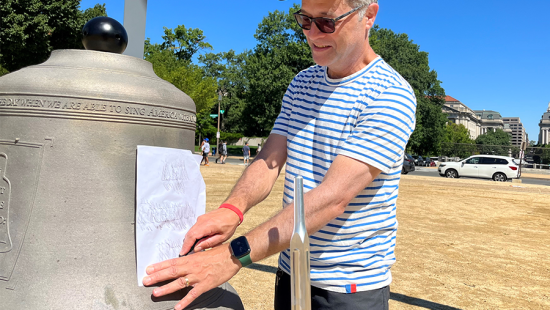 Man wearing a striped shirt and blue baseball hat doing an artistic rubbing of an engraving on a bell