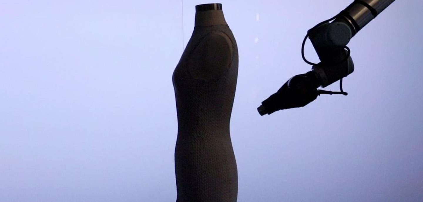 Side-view of a mannequin wearing a piece of fabric being worked by a mechanical arm.