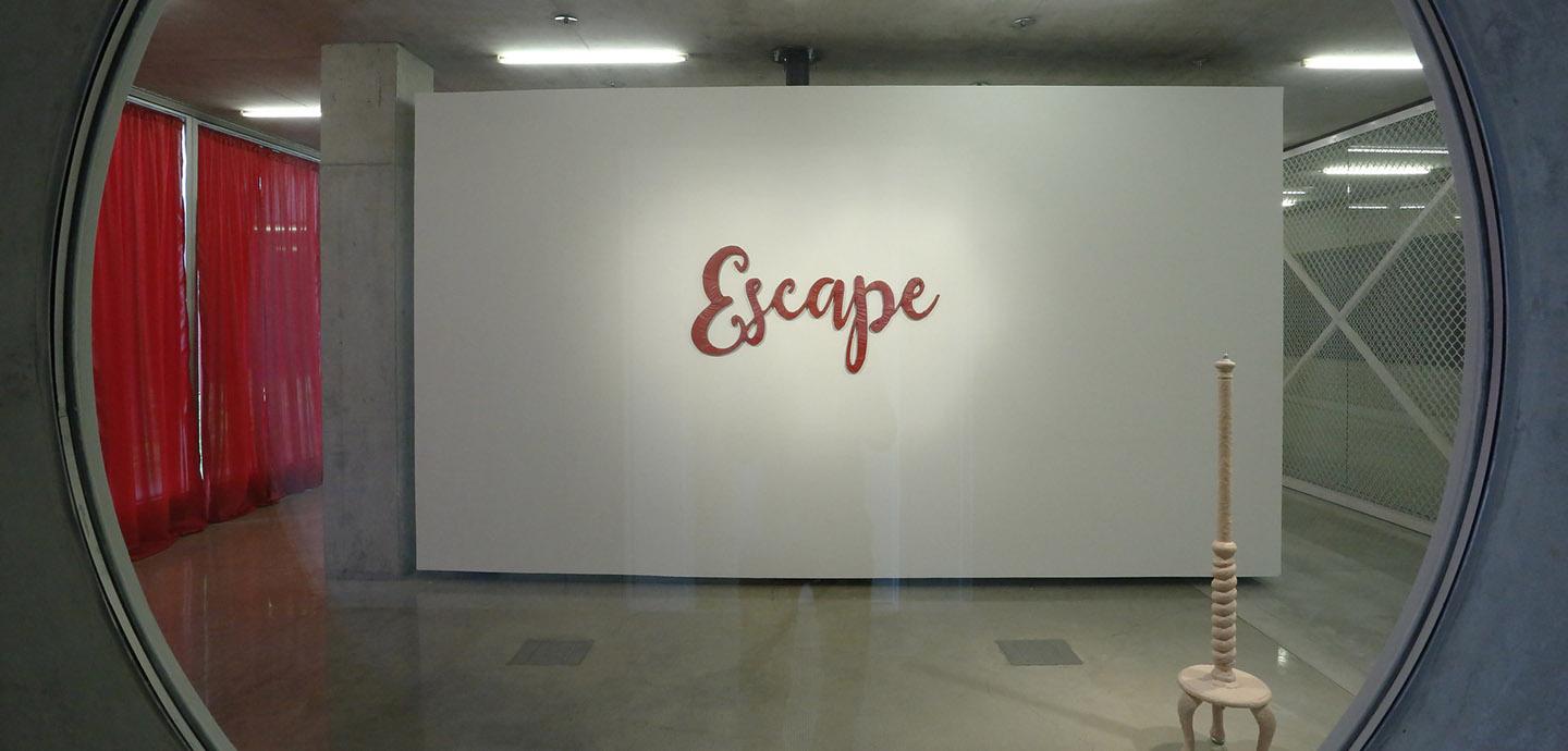 Circle window with an abstract wooden sculpture and the word ESCAPE in red.