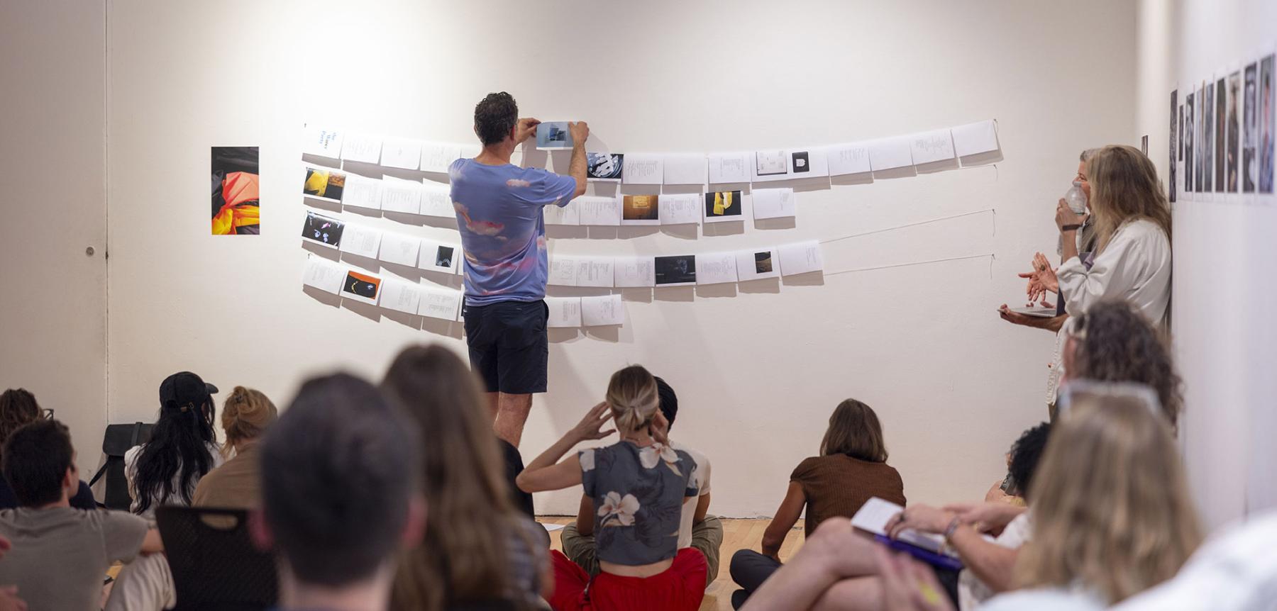 Person hanging work on a wall with an audience of people sitting on the floor behind them.